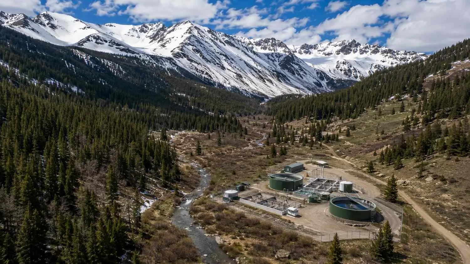 Aerial view of a Colorado mountain watershed showing spring snowpack on high ridgelines above a valley with a small stream and wastewater treatment facility.